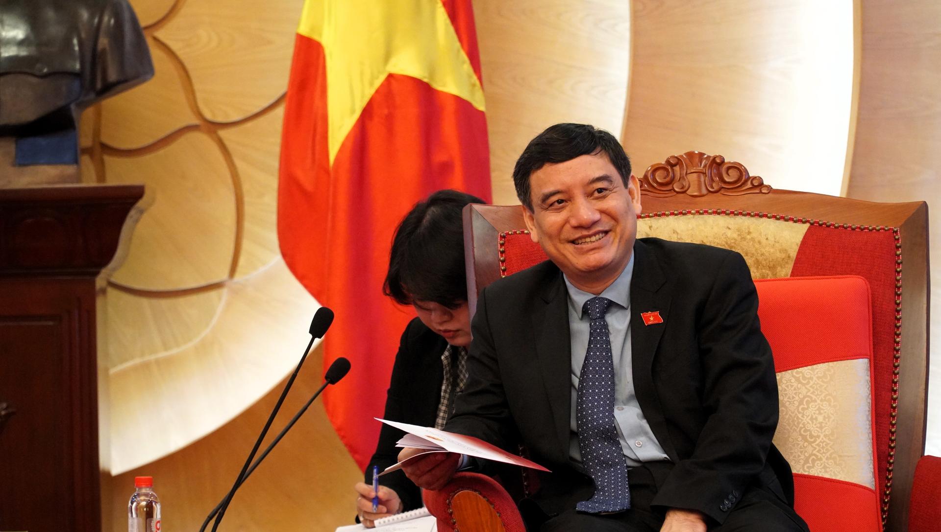 Smiling man in a suit sits on a red-cushioned sofa beside a woman who is reading a notebook; flag in the background.