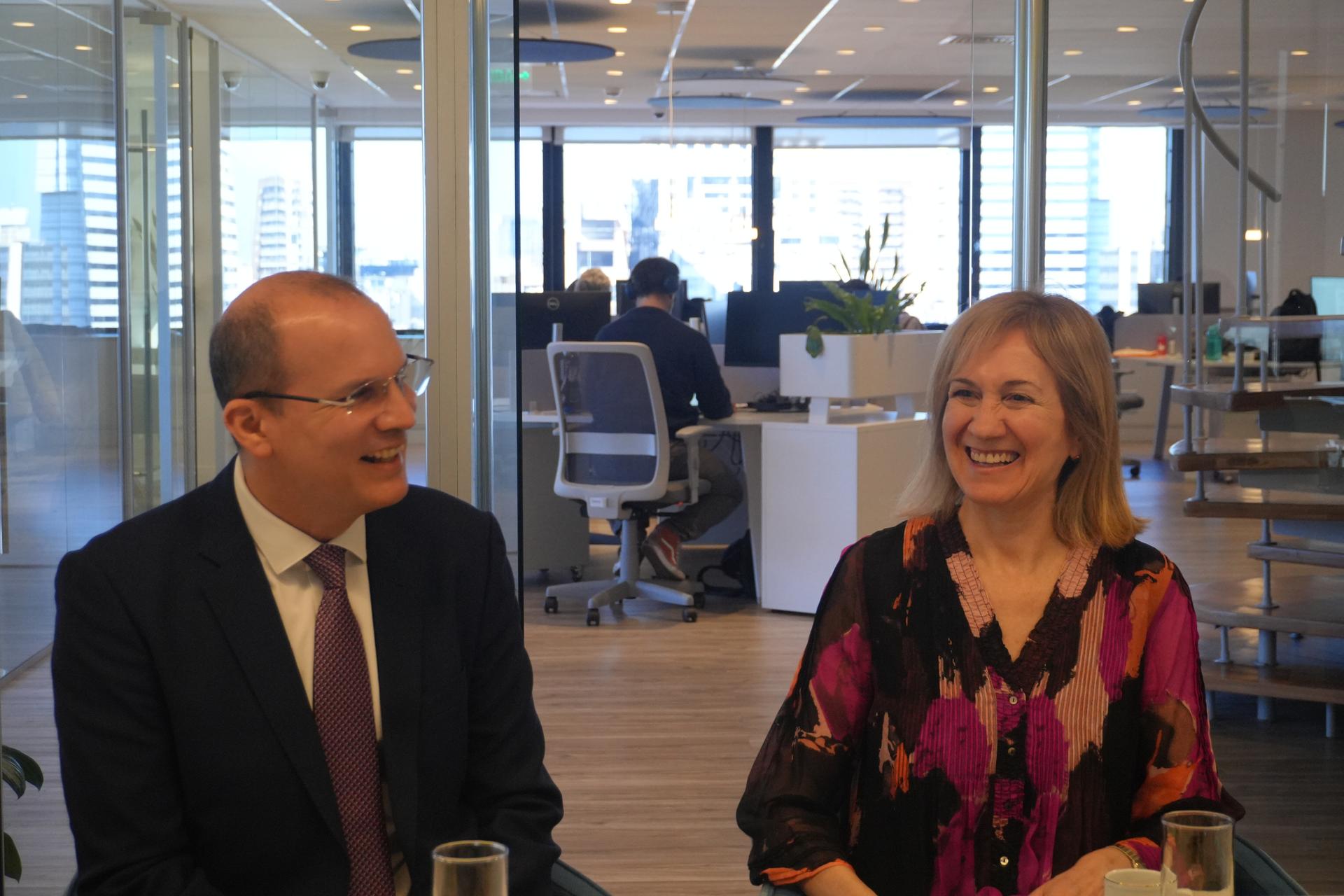 Photo: man in a suit and woman in a floral blouse seated at a desk in a bright office.