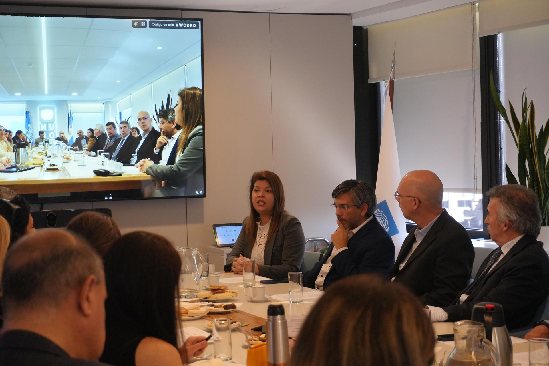 Photograph of a formal business meeting around a conference table; remote participants on screen.
