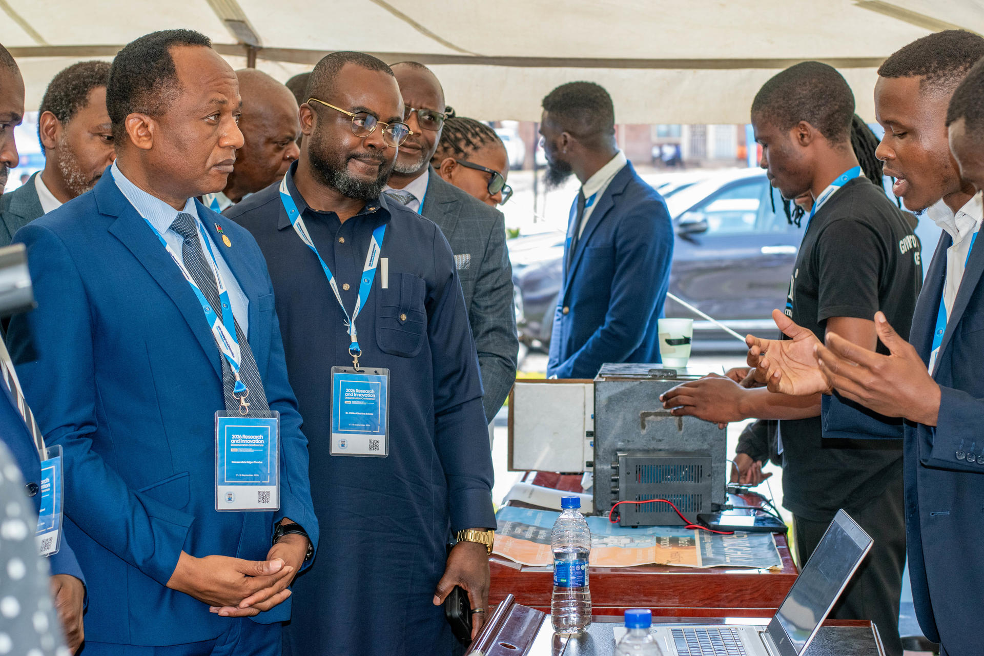 Group of men in blue blazers at a conference booth, examining a device on a table.