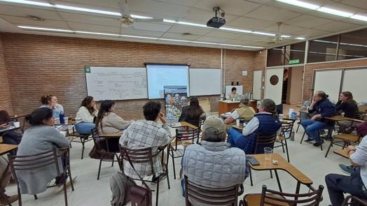 Group of adults seated in a brick-walled classroom listening to a presentation at the front.