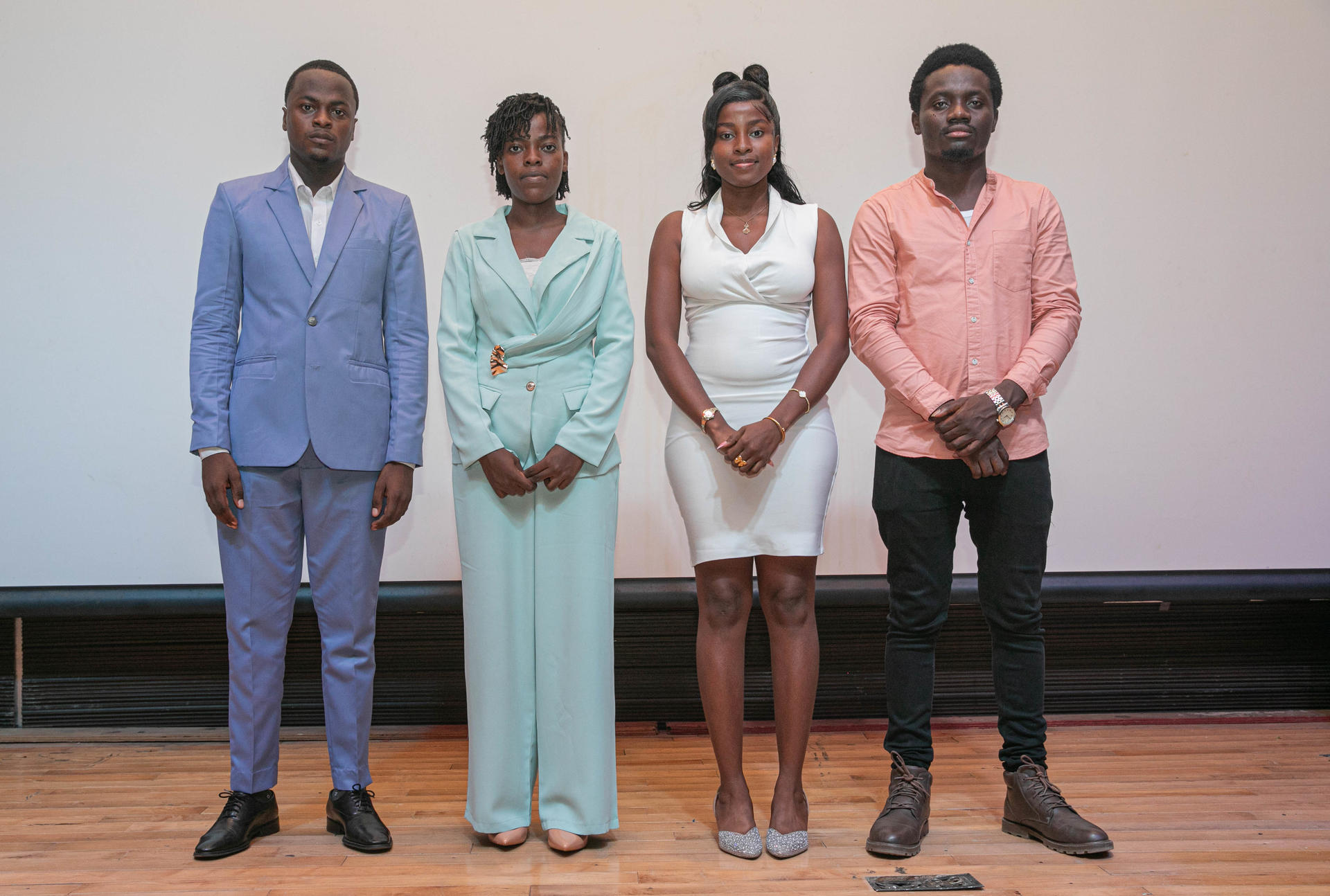 Four adults on stage: man in blue suit, woman in teal dress, woman in white, man in pink shirt