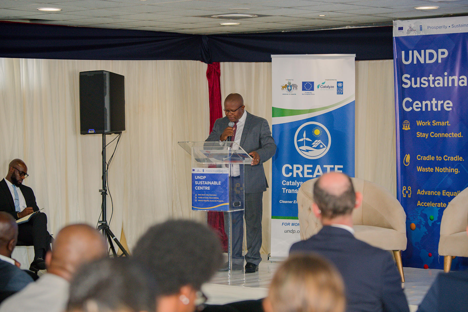 Speaker at a podium in a tent; audience seated, blue UNDP banners in the background.