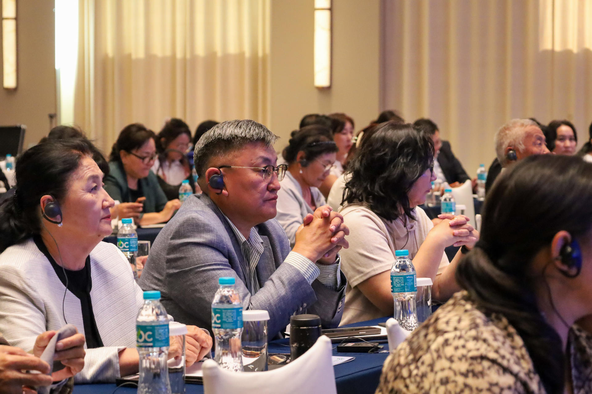 Photograph of an audience at a conference hall listening attentively; water bottles on tables.