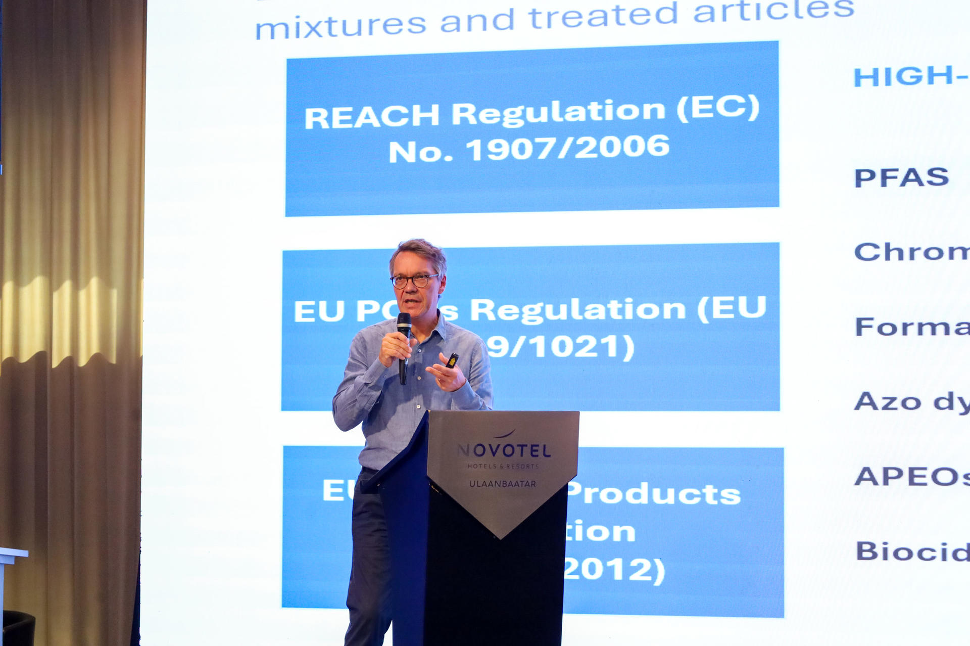 Male presenter on stage delivering a talk with a blue slide deck behind him.