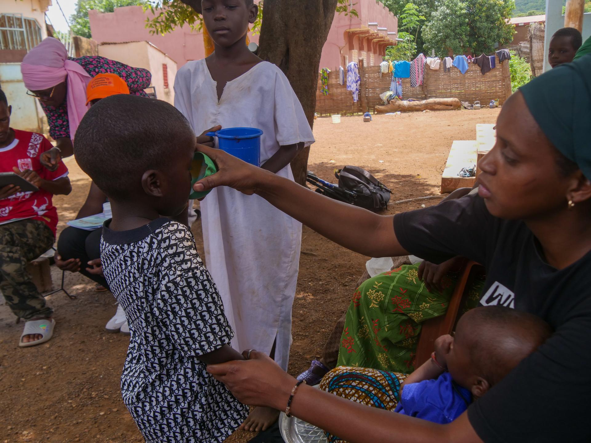 Medical worker vaccinates a child as villagers watch in a dusty village.