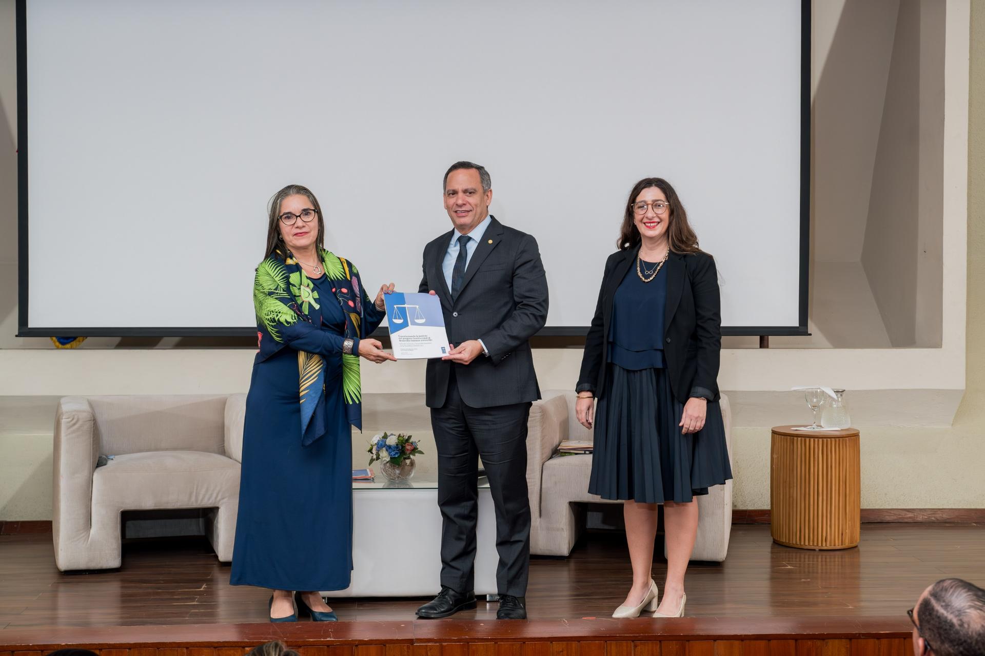 Four people on stage at an award ceremony; man in suit hands certificate to woman in blue dress.