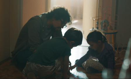 Un adulto con gafas interactúa con dos niños pequeños en el suelo de una habitación iluminada por luz natural. Los niños están concentrados en una actividad con piezas o juguetes en el suelo. Al fondo, se observa un juguete de madera, destacando un ambiente cálido y familiar
