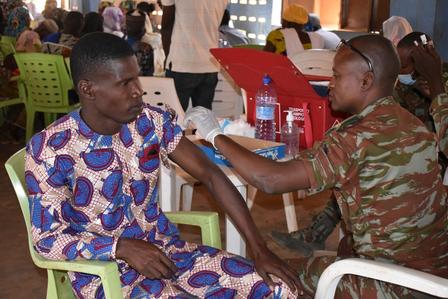 A soldier administers a vaccine to a young man sitting in a chair.