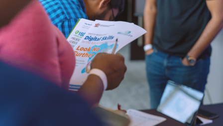 A person holds papers while standing near a table, with another person in a blue shirt nearby.