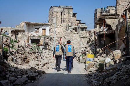Two aid workers walking through a demolished urban area with rubble and damaged buildings.