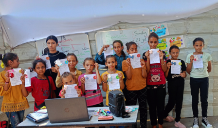 Group of children holding drawings in a classroom setting with a laptop on a table.