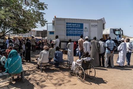 A crowd of people gathers around a mobile medical truck in a sunny outdoor setting.
