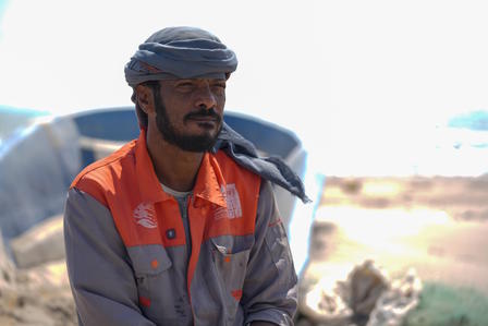 Man in work attire sitting near a boat, with a calm expression and a coastal background.