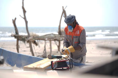 A worker in safety gear repairs a boat on a beach with waves in the background.