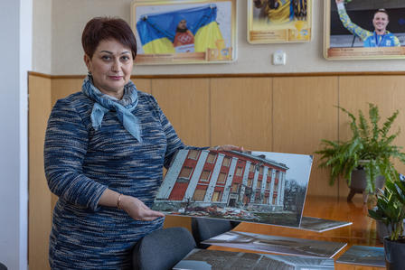 A woman in a blue sweater holds a photo of a building, surrounded by plants and framed images.