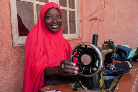 Smiling woman in red hijab holding a sewing machine wheel in a brightly colored room.