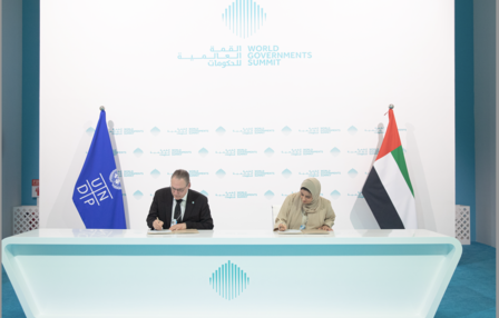 Two officials seated at a table, signing documents with flags in the background.
