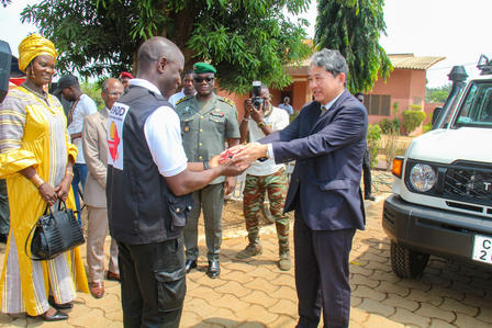 A man in formal attire hands over an item to another man in a vest, surrounded by onlookers.