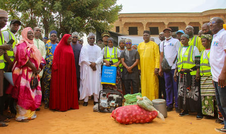 A diverse group of people in colorful clothing gathers outdoors, with tools and produce displayed.
