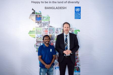 Two men stand in front of a colorful map of Bangladesh, smiling.