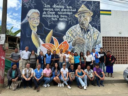 A diverse group of people poses in front of a colorful mural depicting two elderly figures.