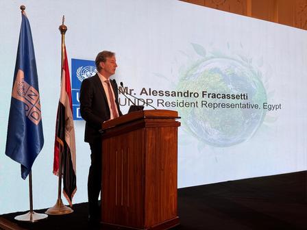 A man in a suit speaks at a podium, flanked by flags, in a conference setting.