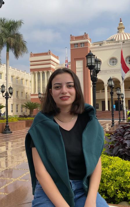 A young woman with a green shawl smiles in front of an ornate building and palm trees.