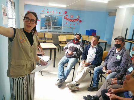 A woman gestures while presenting to a group of seated men in a classroom setting.