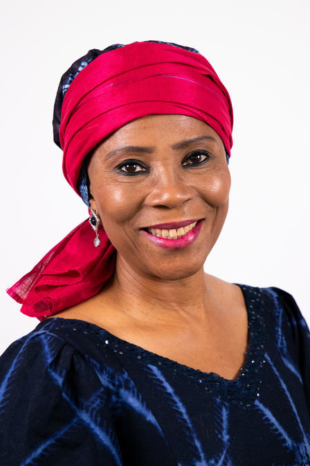Smiling woman wearing a bright pink headwrap and dark patterned clothing against a white background.