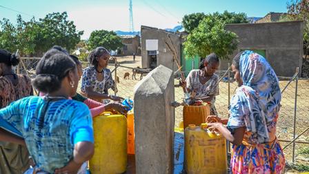 Women gathering water from a pump, with colorful containers in an outdoor setting.