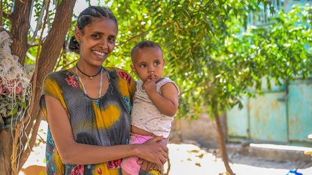Smiling woman holding a baby against a leafy outdoor background.