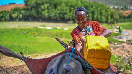 A woman smiling while filling a yellow container with water from a donkey.