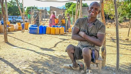 A smiling man seated on a chair, surrounded by colorful water containers and hills in the background.