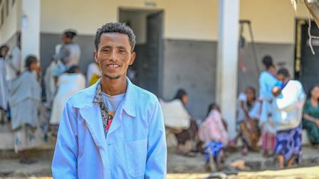 Young man in a blue lab coat smiles, with a busy outdoor scene of people in the background.