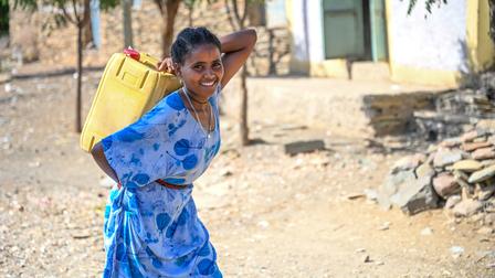 A smiling girl in a blue dress carries a yellow water container along a dirt path.