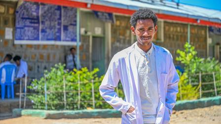 A smiling healthcare worker in a white coat stands outdoors in front of a clinic.