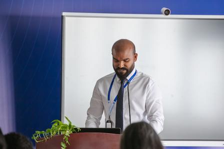 A man in a white shirt and tie stands at a podium, speaking to an audience.