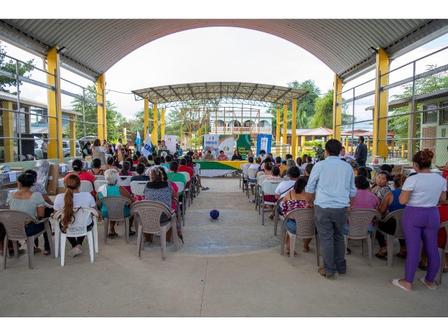 A large crowd seated in chairs under a roofed area with a stage in the background.