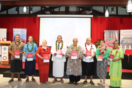Nine individuals stand together on stage, holding certificates and wearing traditional attire.