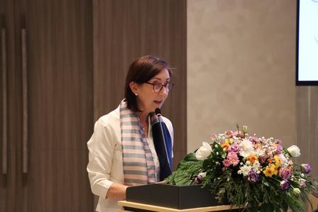 A woman speaking at a podium with a floral arrangement, dressed in a white jacket and scarf.