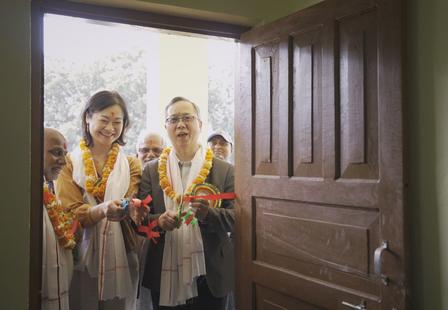 A group of smiling people with flower garlands stand by a doorway, preparing to cut a ribbon.