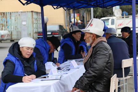 People in blue jackets seated at tables, interacting outdoors in a busy urban setting.