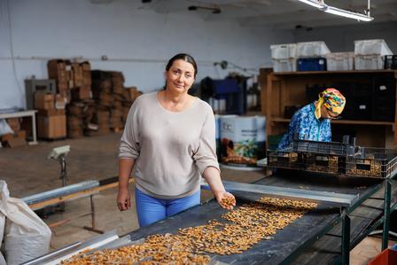 A woman smiles while sorting nuts on a conveyor belt in a warehouse. Another worker is nearby.