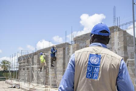 A man wearing a UNDP vest observes workers constructing a building under a clear blue sky.