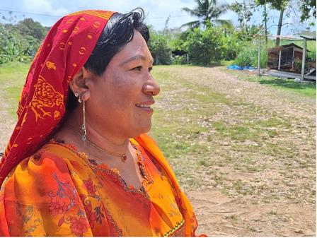 A woman in colorful attire and a red scarf smiles, set against a green, open landscape.