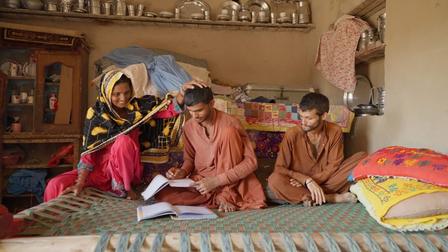 A woman and two boys study together in a simple room filled with household items.