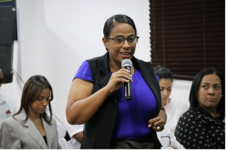 A woman in a blue top speaks into a microphone while seated attendees listen.
