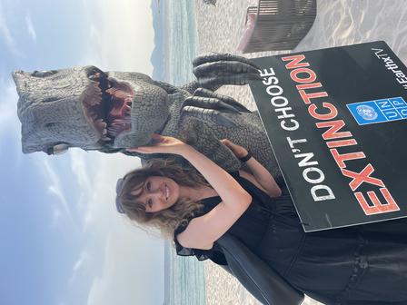 A woman poses with a dinosaur costume and a protest sign on a beach.