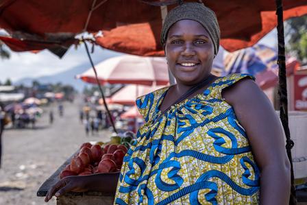 Smiling woman in colorful attire stands at a market stall with tomatoes.